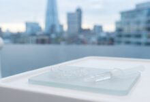 A photorealistic image of a professional teeth whitening kit with custom trays and a gel syringe on a sterile surface inside a modern dental clinic, with a blurred view of the London, UK skyline in the background.