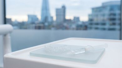 A photorealistic image of a professional teeth whitening kit with custom trays and a gel syringe on a sterile surface inside a modern dental clinic, with a blurred view of the London, UK skyline in the background.