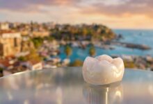 A close-up, photorealistic image of a perfect white dental crown resting on a surface, with the beautiful, blurred coastline of a city in Turkey visible in the background during a warm sunset.