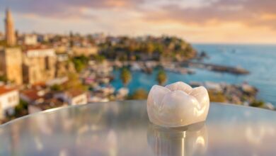 A close-up, photorealistic image of a perfect white dental crown resting on a surface, with the beautiful, blurred coastline of a city in Turkey visible in the background during a warm sunset.