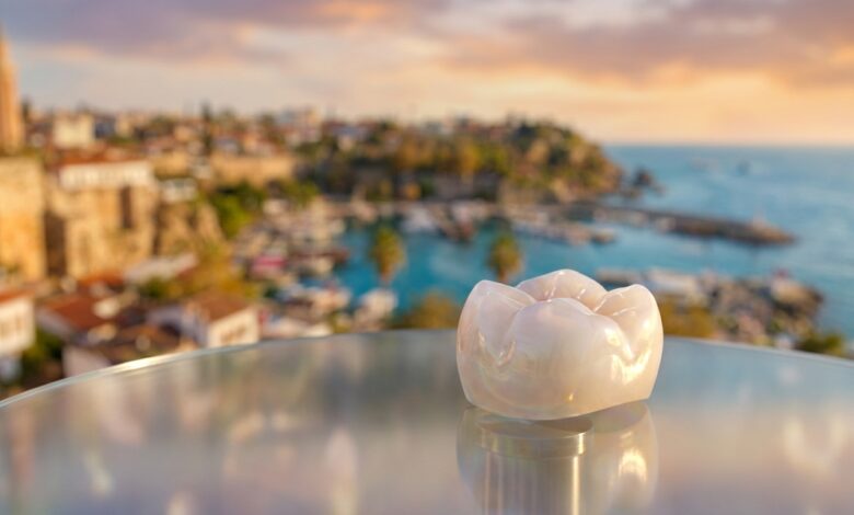 A close-up, photorealistic image of a perfect white dental crown resting on a surface, with the beautiful, blurred coastline of a city in Turkey visible in the background during a warm sunset.