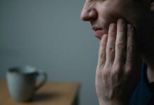 A photorealistic close-up of a person holding their jaw in pain, symbolizing the warning signs of a tooth infection. The lighting is dramatic and the background is blurred.