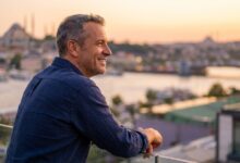 A happy man in his late 40s with a perfect smile stands on a hotel balcony, with a beautifully blurred sunset view of the Istanbul skyline in the background.