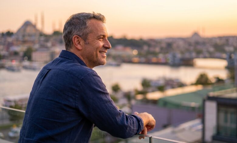 A happy man in his late 40s with a perfect smile stands on a hotel balcony, with a beautifully blurred sunset view of the Istanbul skyline in the background.