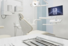 A clean, brightly lit modern dental examination room with a white dental chair, an overhead operating light, and blurred dental instruments on a tray in the foreground. Advanced, sleek dental equipment is visible in the background, conveying a sense of high quality and professionalism.