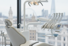 A photorealistic image of a sophisticated, modern dental treatment room in the UK. The scene features a pristine, empty ergonomic dental chair, sleek advanced dental instruments, and a soft, bright clinical light. Through a large window in the background, a very softly blurred London cityscape is visible. The image conveys a sense of cleanliness, high technology, and patient comfort, with no visible text.