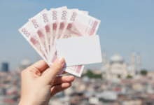 A person's hand holding blurred UK pound notes or a generic credit card, with a stunning, sunlit and slightly blurred panoramic view of the Istanbul skyline, featuring minarets and historic architecture, under a clear blue sky in the background.