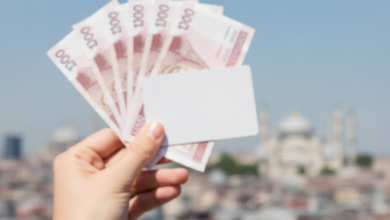 A person's hand holding blurred UK pound notes or a generic credit card, with a stunning, sunlit and slightly blurred panoramic view of the Istanbul skyline, featuring minarets and historic architecture, under a clear blue sky in the background.
