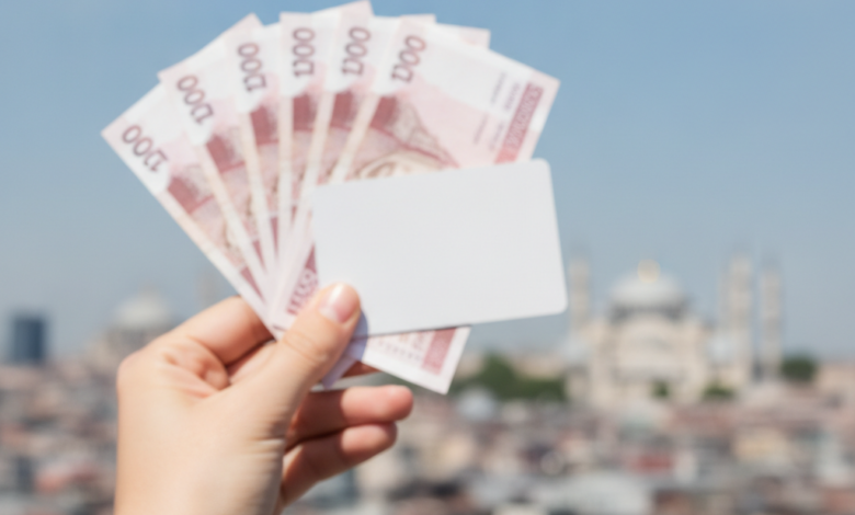 A person's hand holding blurred UK pound notes or a generic credit card, with a stunning, sunlit and slightly blurred panoramic view of the Istanbul skyline, featuring minarets and historic architecture, under a clear blue sky in the background.